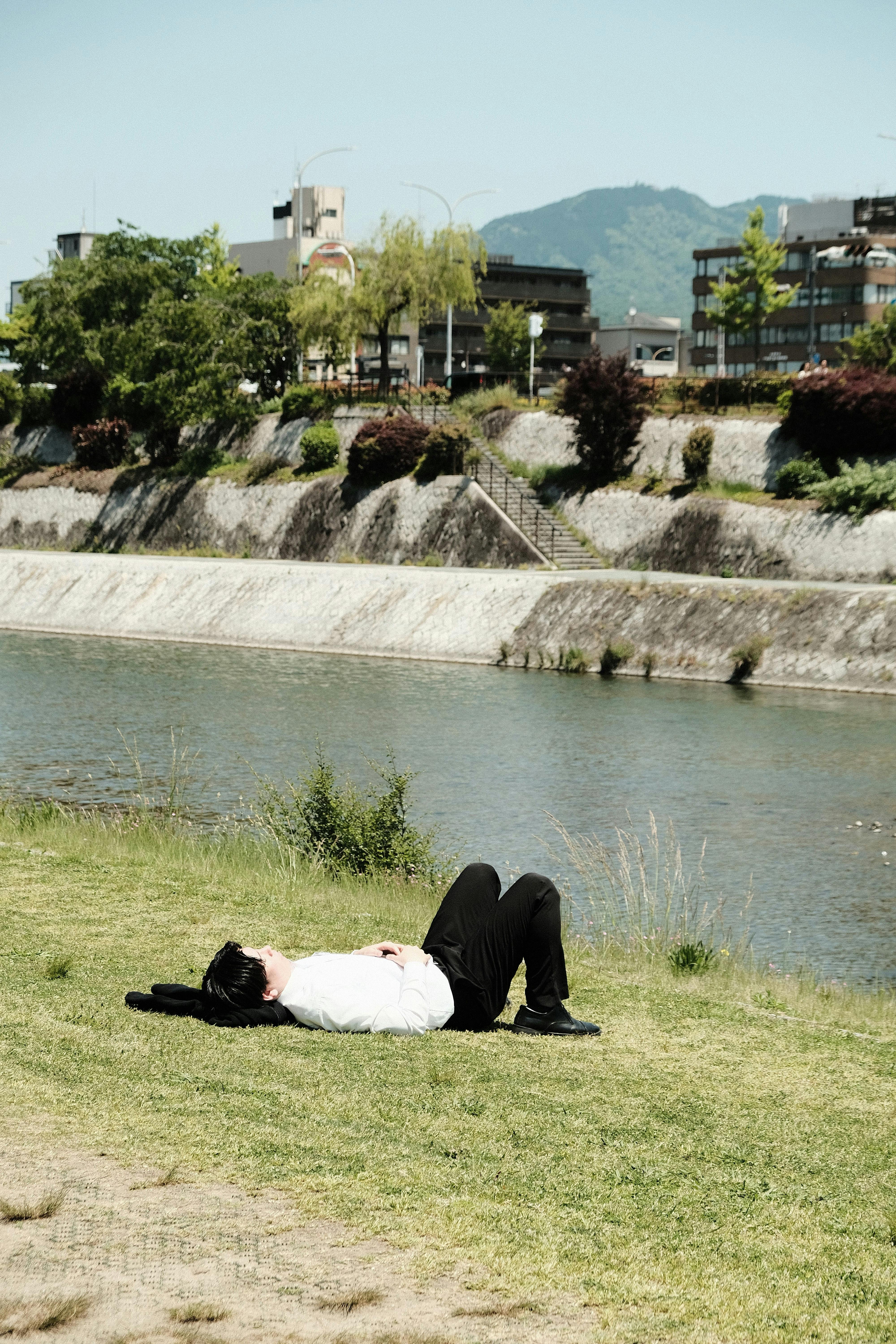 Person lying on grass by a river in a sunny urban park area.