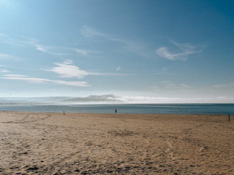 Serene beach landscape with a solitary figure under a vast blue sky and gentle waves.