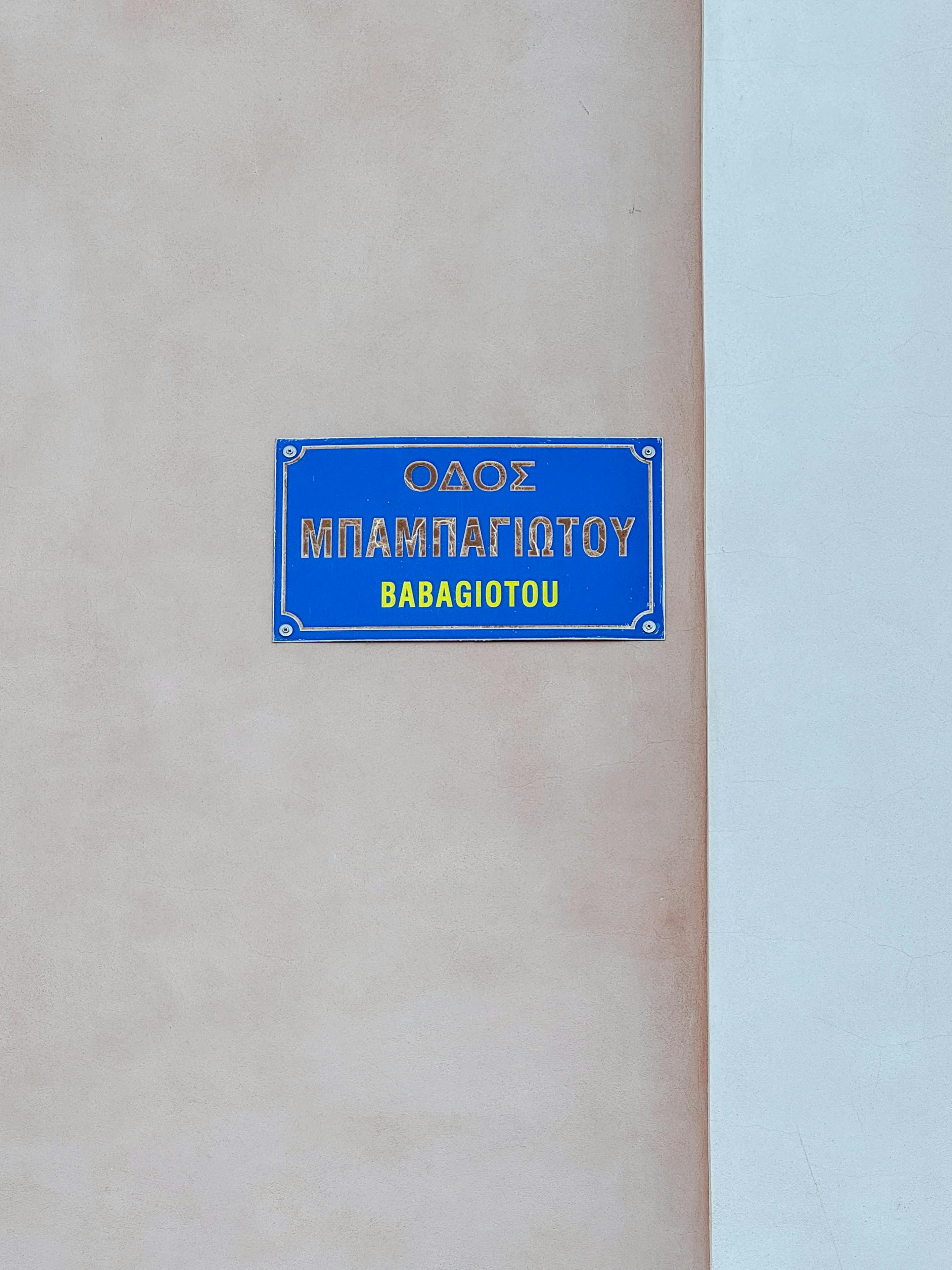 Gratis Cartello stradale con la scritta greca 'Babagiotou' su un muro color pastello in Grecia. Foto a disposizione