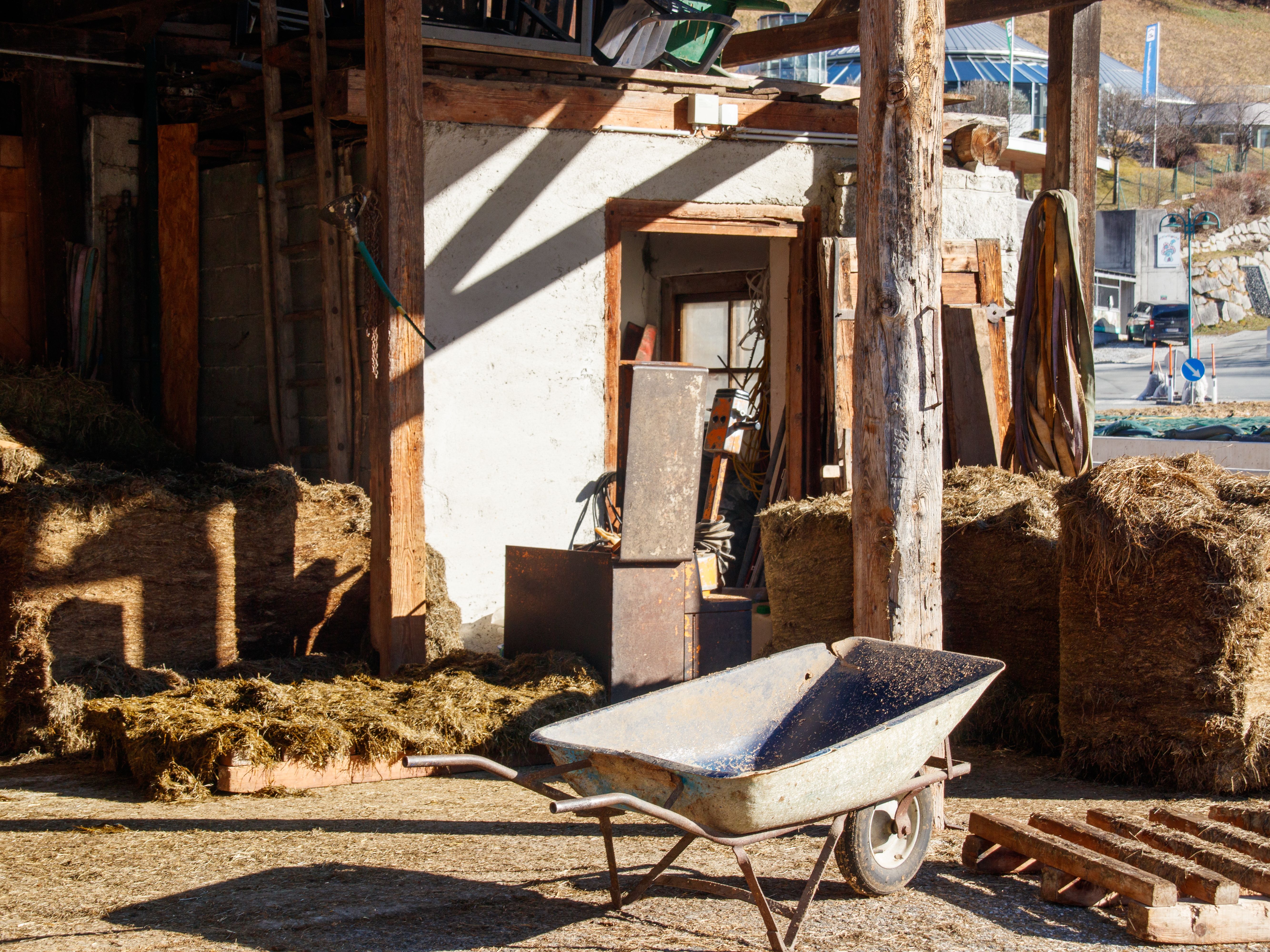 A rural farmyard scene featuring a wheelbarrow, hay bales, and wooden structures under sunlight.