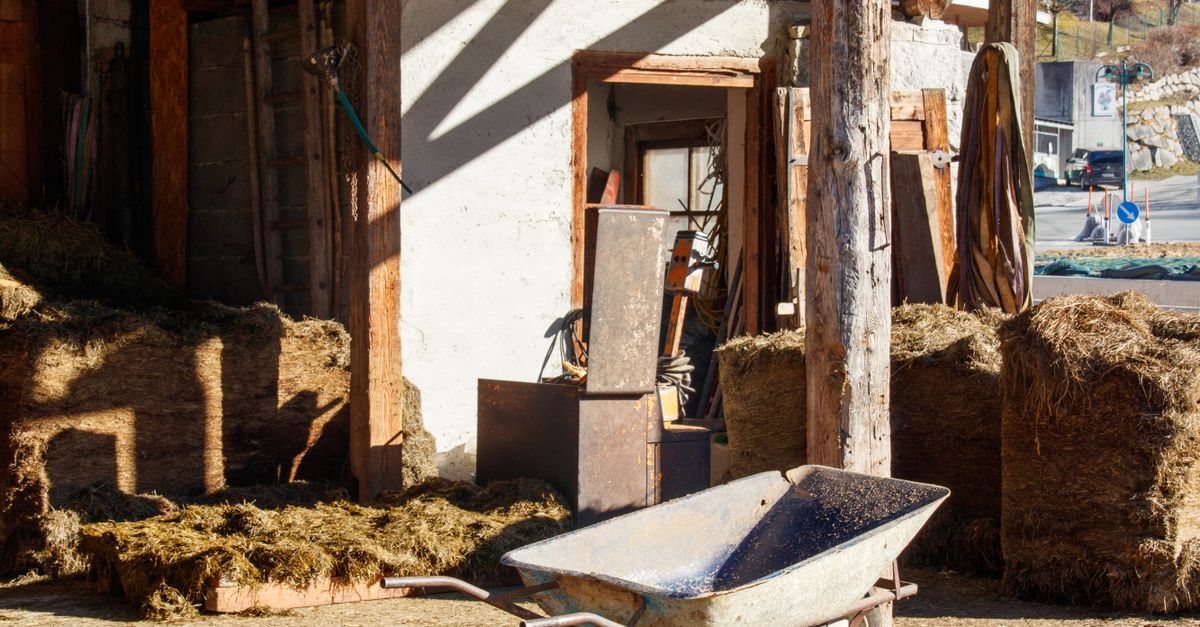 A rural farmyard scene featuring a wheelbarrow, hay bales, and wooden structures under sunlight.