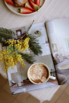 A cozy scene featuring a cappuccino, magazine, and mimosa flowers with strawberries.
