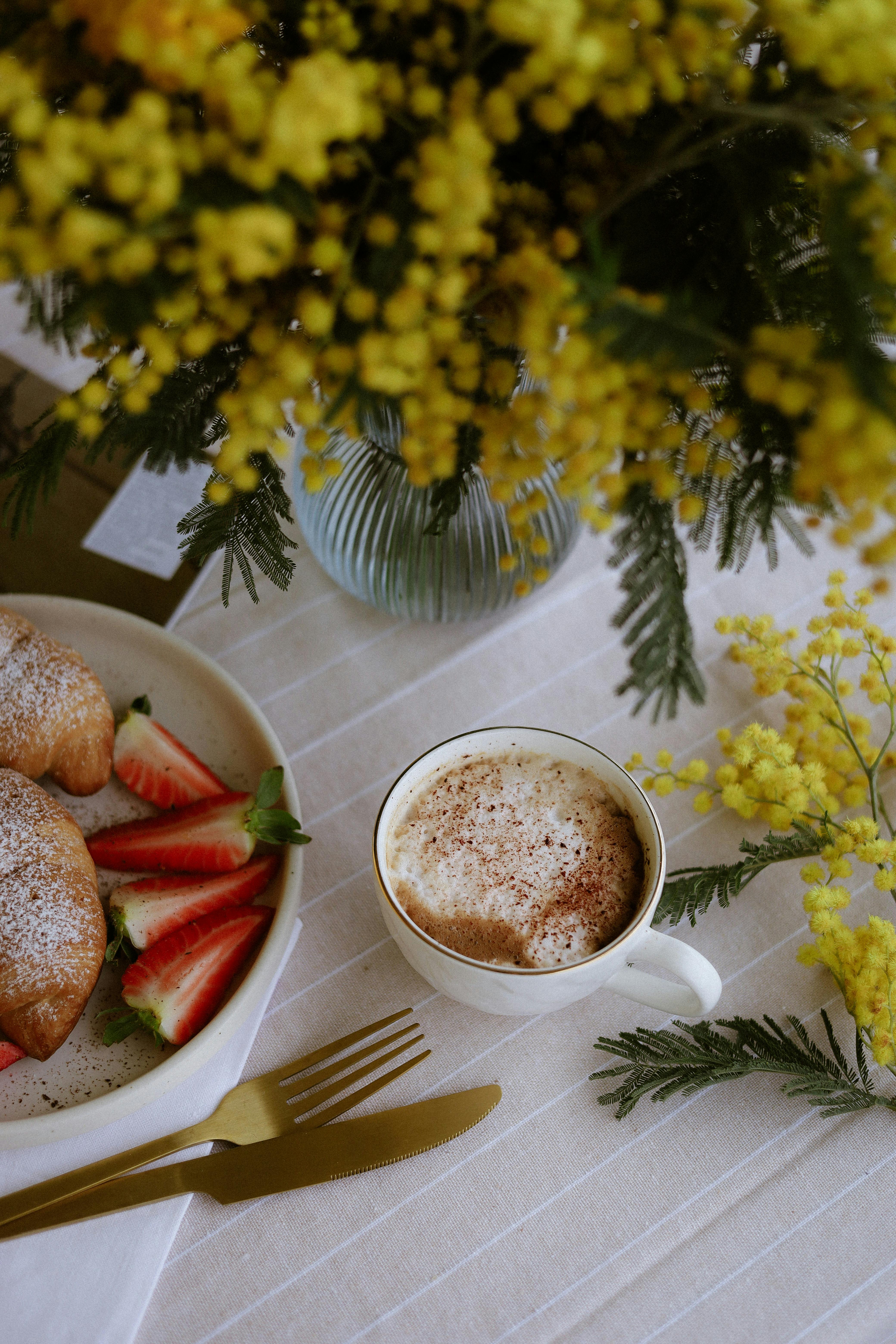 A cozy breakfast setting with cappuccino, croissants, strawberries, and vibrant yellow flowers.