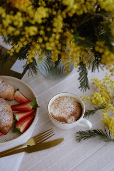 A cozy breakfast setting with cappuccino, croissants, strawberries, and vibrant yellow flowers.