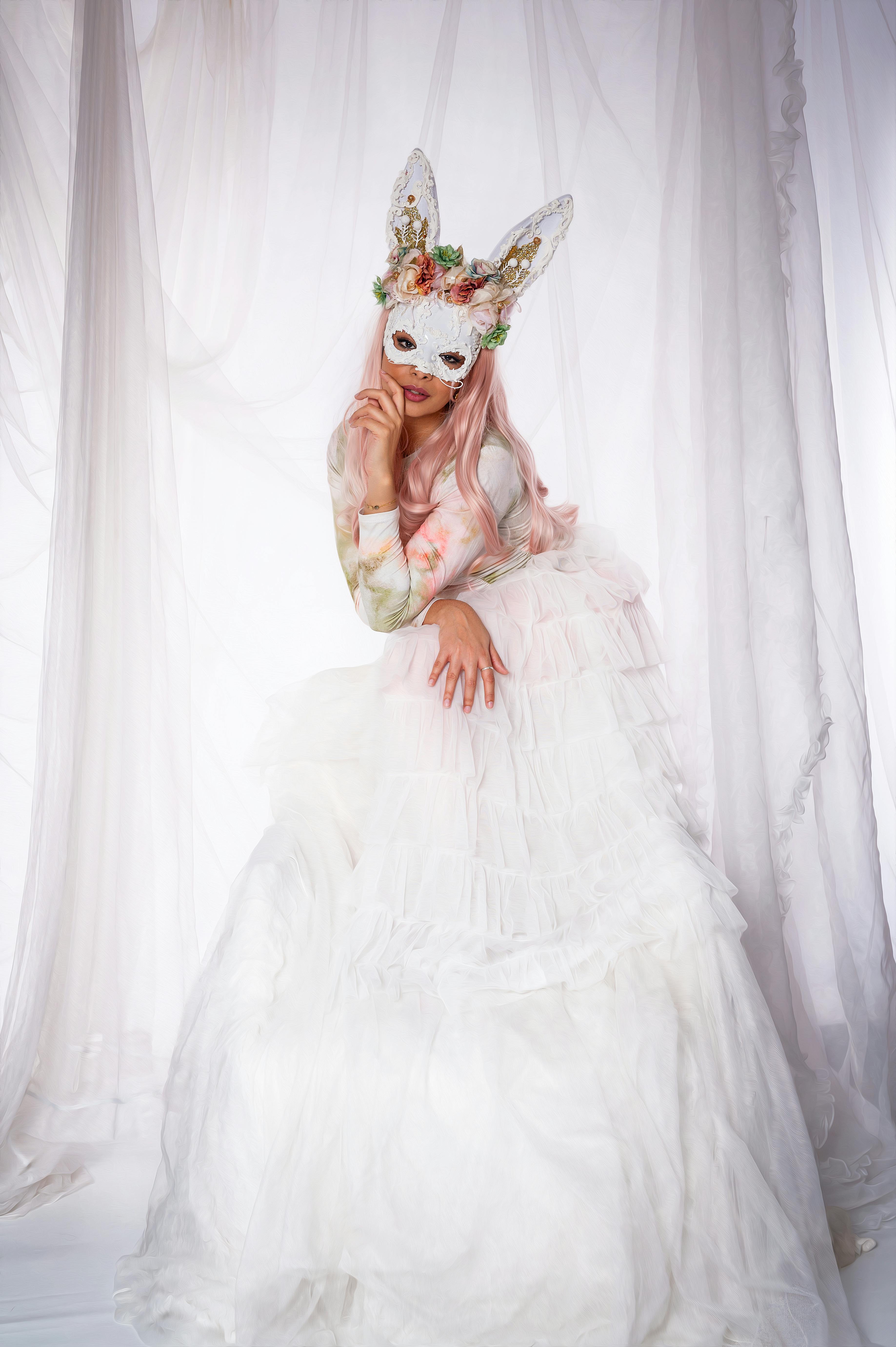 Elegant woman in a white gown and floral mask posing among sheer curtains.