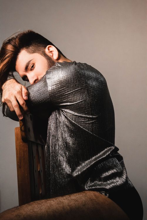 Free A fashionable man in a metallic shirt poses stylishly on a chair indoors, showcasing modern fashion trends. Stock Photo