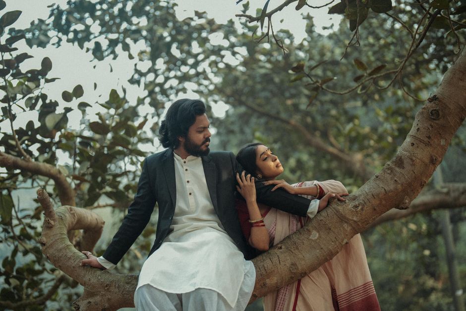 A serene moment with a couple in traditional attire sitting on a large tree branch in a lush outdoor setting.
