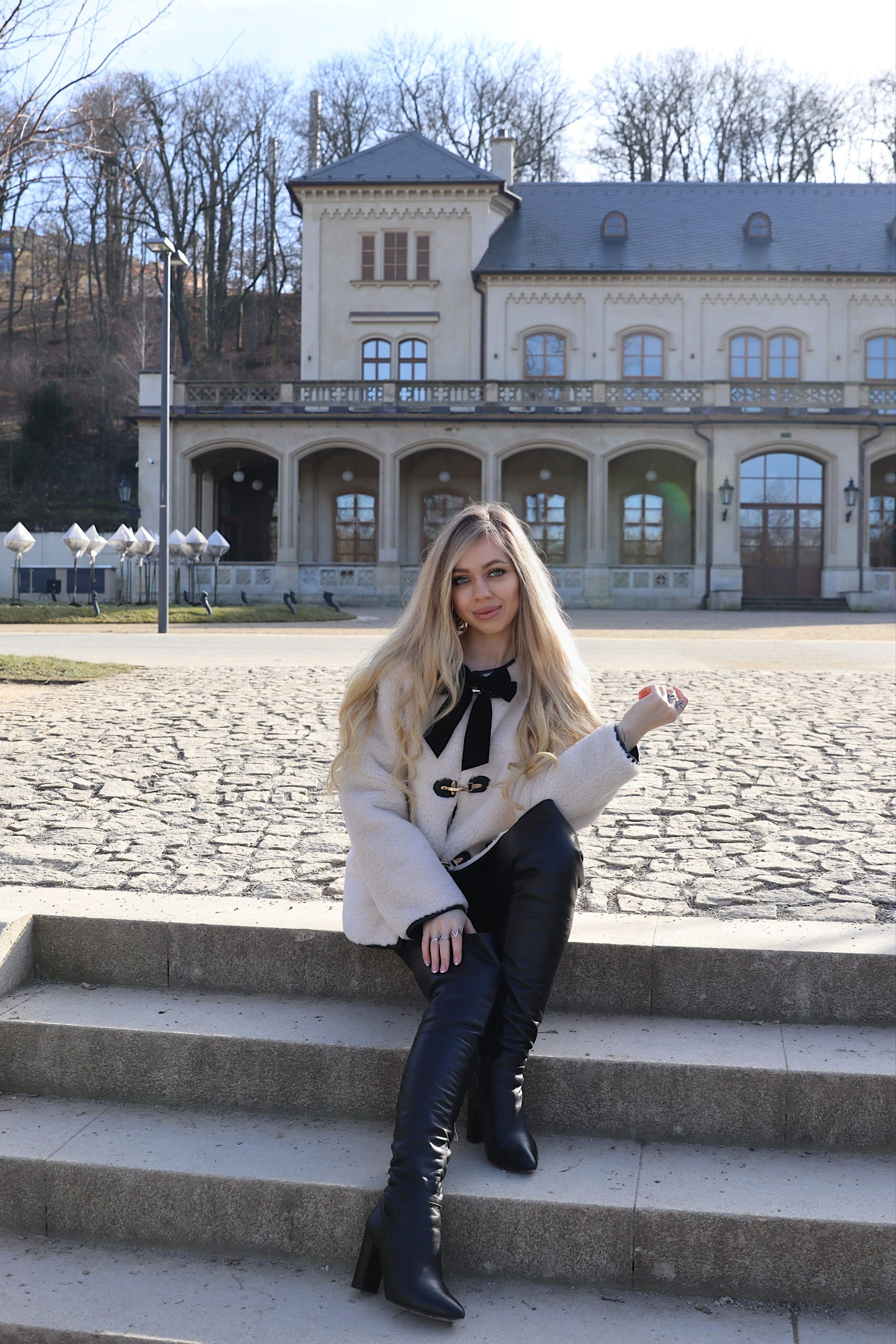 Fashionable woman in winter attire poses on steps before an elegant historical building.