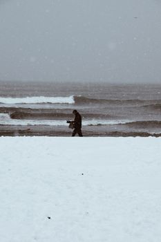 A person walks along a snowy beach with waves crashing in the background during a winter day.