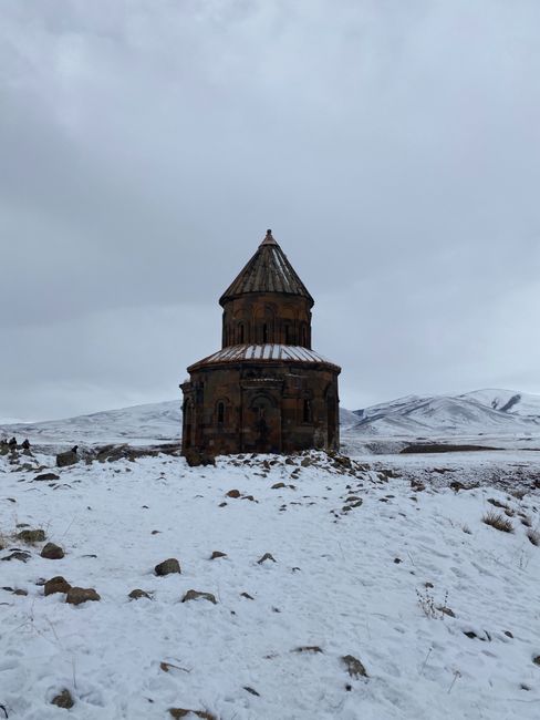 A historic church stands solitary in a serene snowy field against a mountainous backdrop in Ani, Turkey.