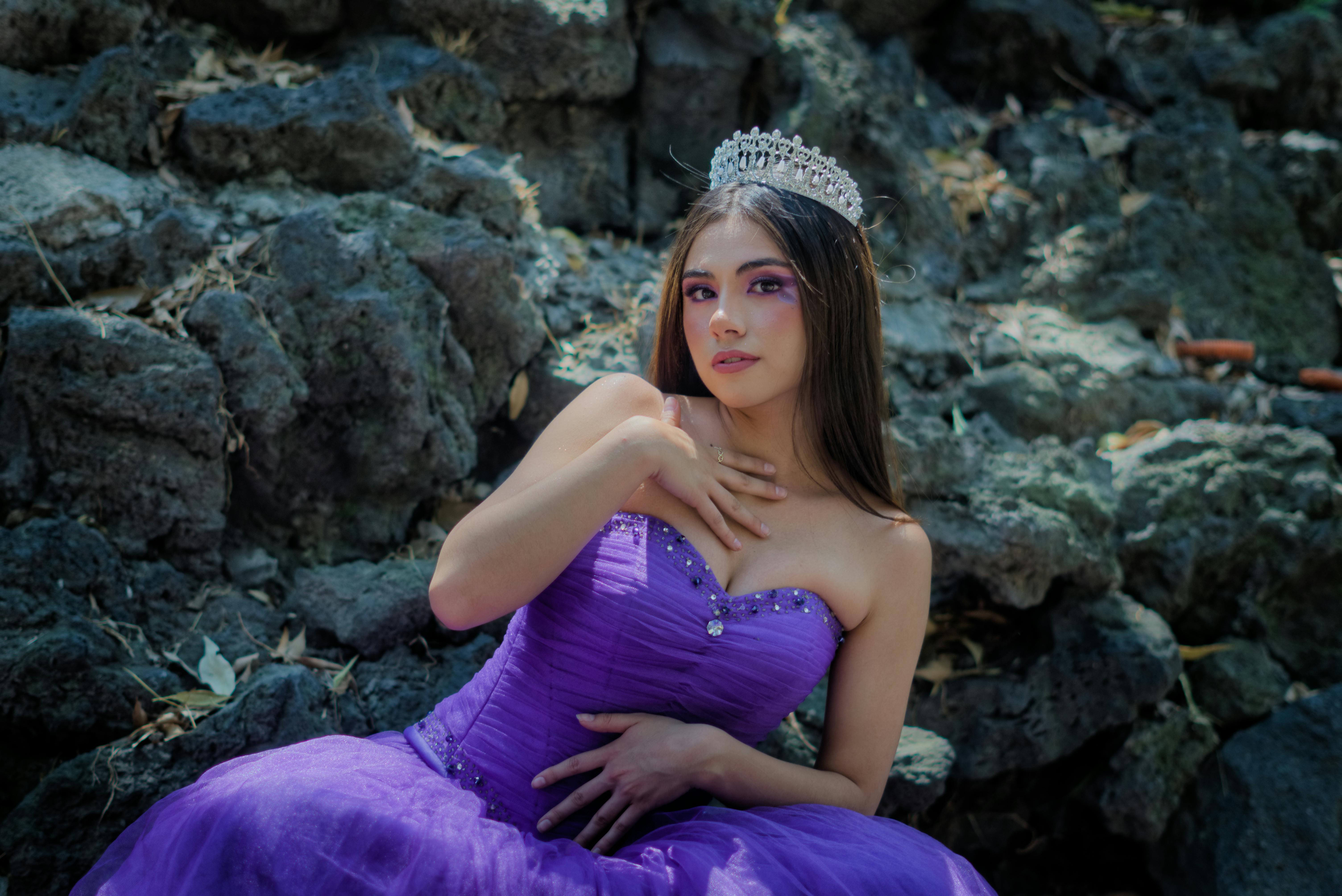 Stunning woman in a purple gown with a tiara posing elegantly outdoors against rocky background.