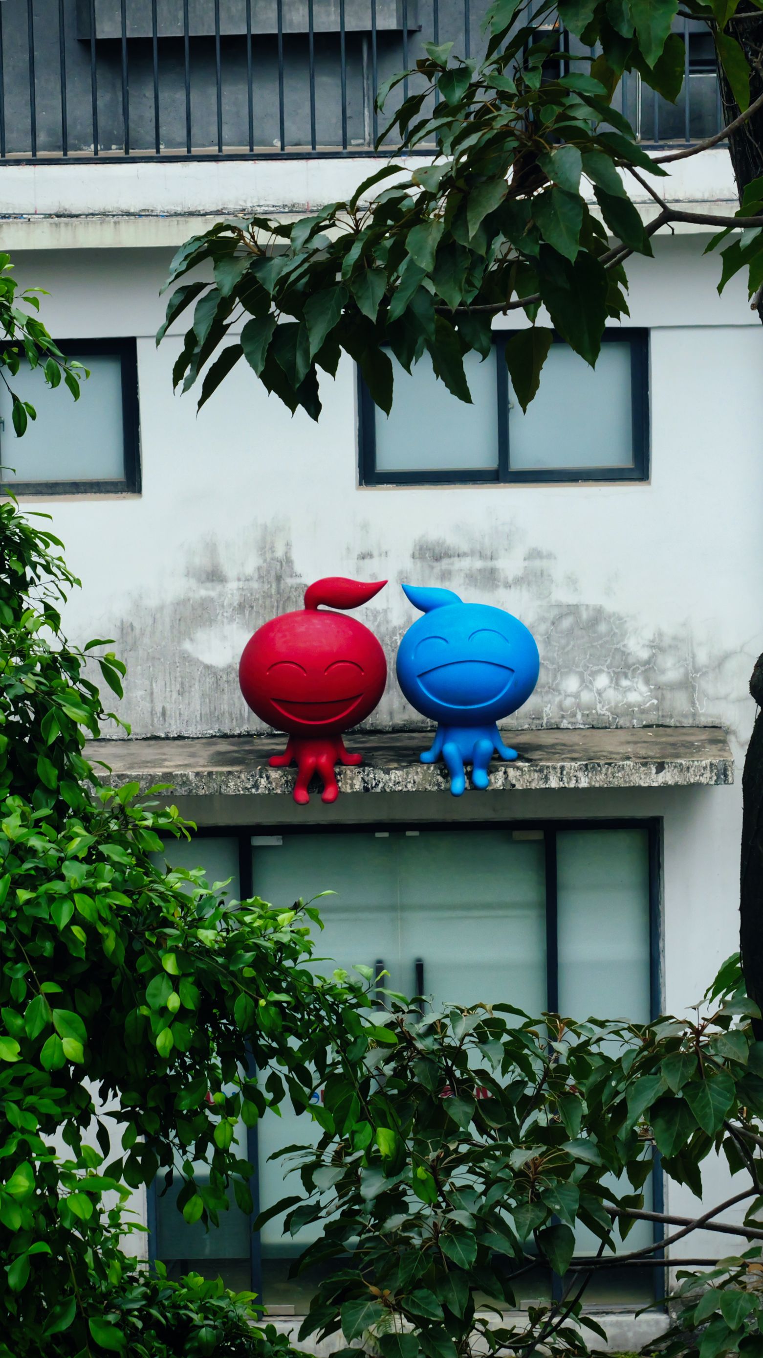 Two vibrant cartoon statues sit on a building ledge amidst greenery.