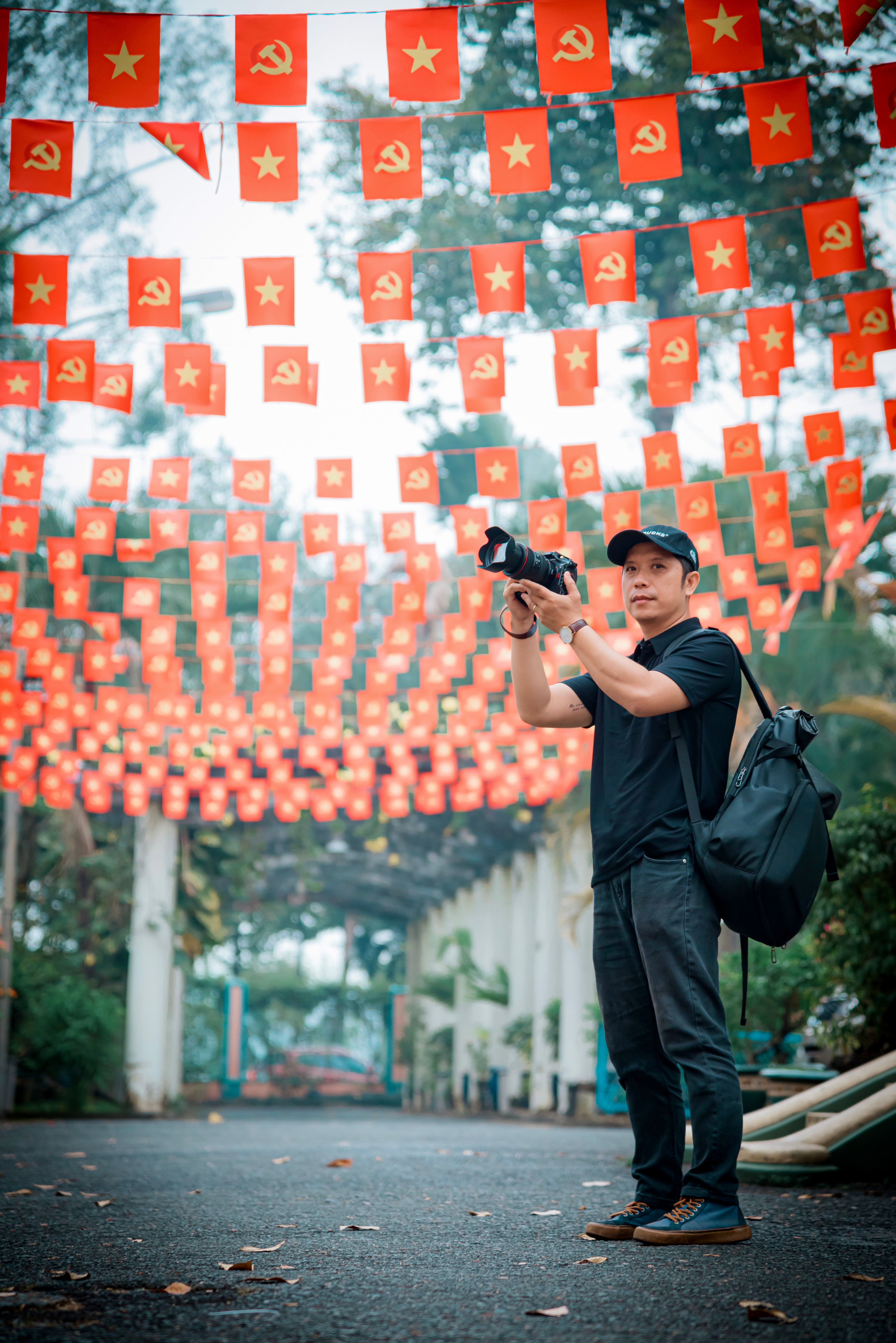 A man with a camera stands under vibrant red flags in a festive outdoor setting.