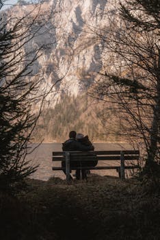 A couple embraces on a bench overlooking a serene mountain lake, surrounded by nature.