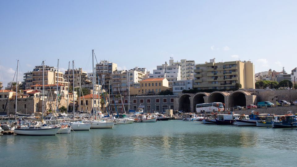 Scenic view of Heraklion marina with yachts and city buildings under a clear blue sky.