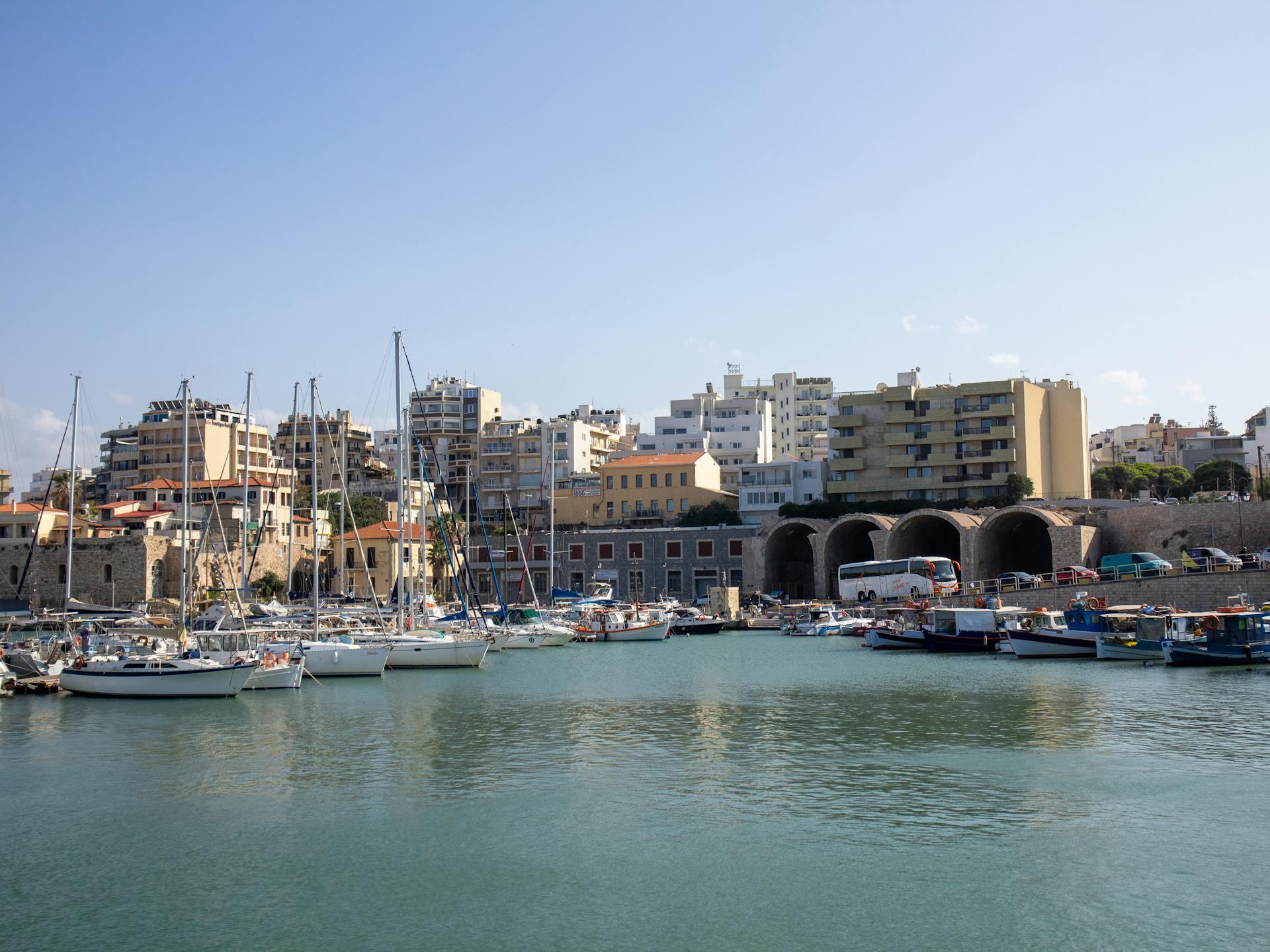 Scenic view of Heraklion marina with yachts and city buildings under a clear blue sky.