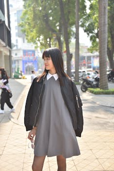 Young woman in a black jacket and gray dress walks confidently in the city.