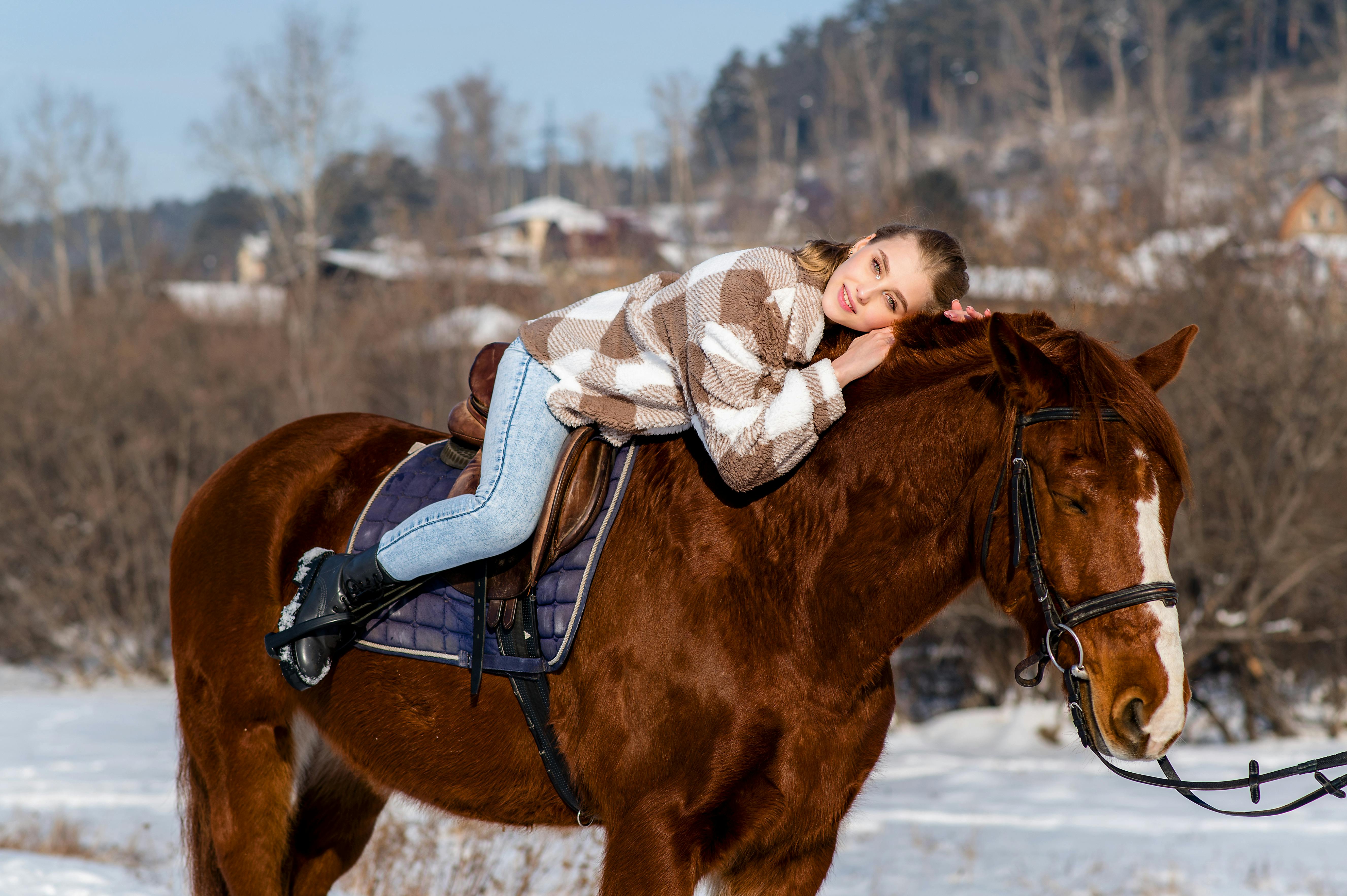 A young woman in a cozy jacket resting on a brown horse in a snowy outdoor setting.