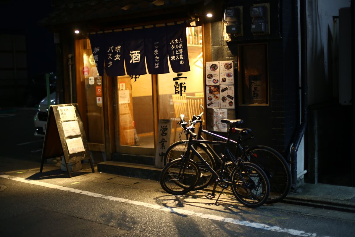 A small izakaya at night in Fukuoka with noren curtains advertising sake, goma-saba and mentaiko pasta