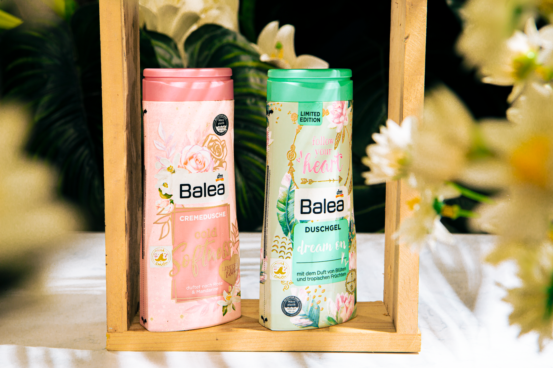 Two Balea shower gels with floral designs displayed in a wooden frame surrounded by flowers.