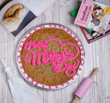 A delicious cookie cake with 'Happy Mother's Day' icing surrounded by baking tools and cookbooks.