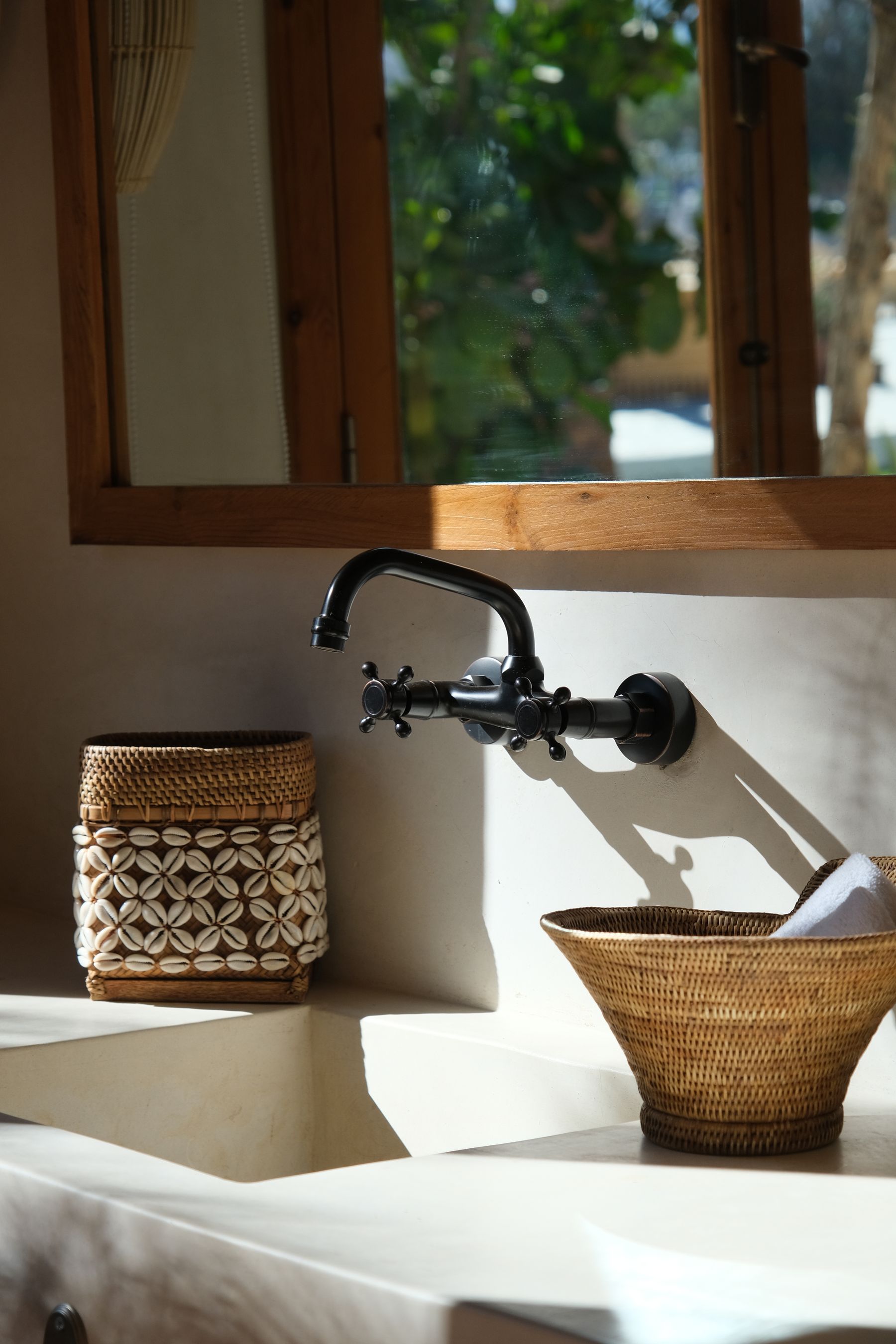 Sunlit bathroom detail with a white basin, dark faucet, timber window and woven containers