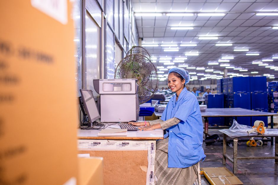 A cheerful female worker at a computer station in a busy warehouse environment.