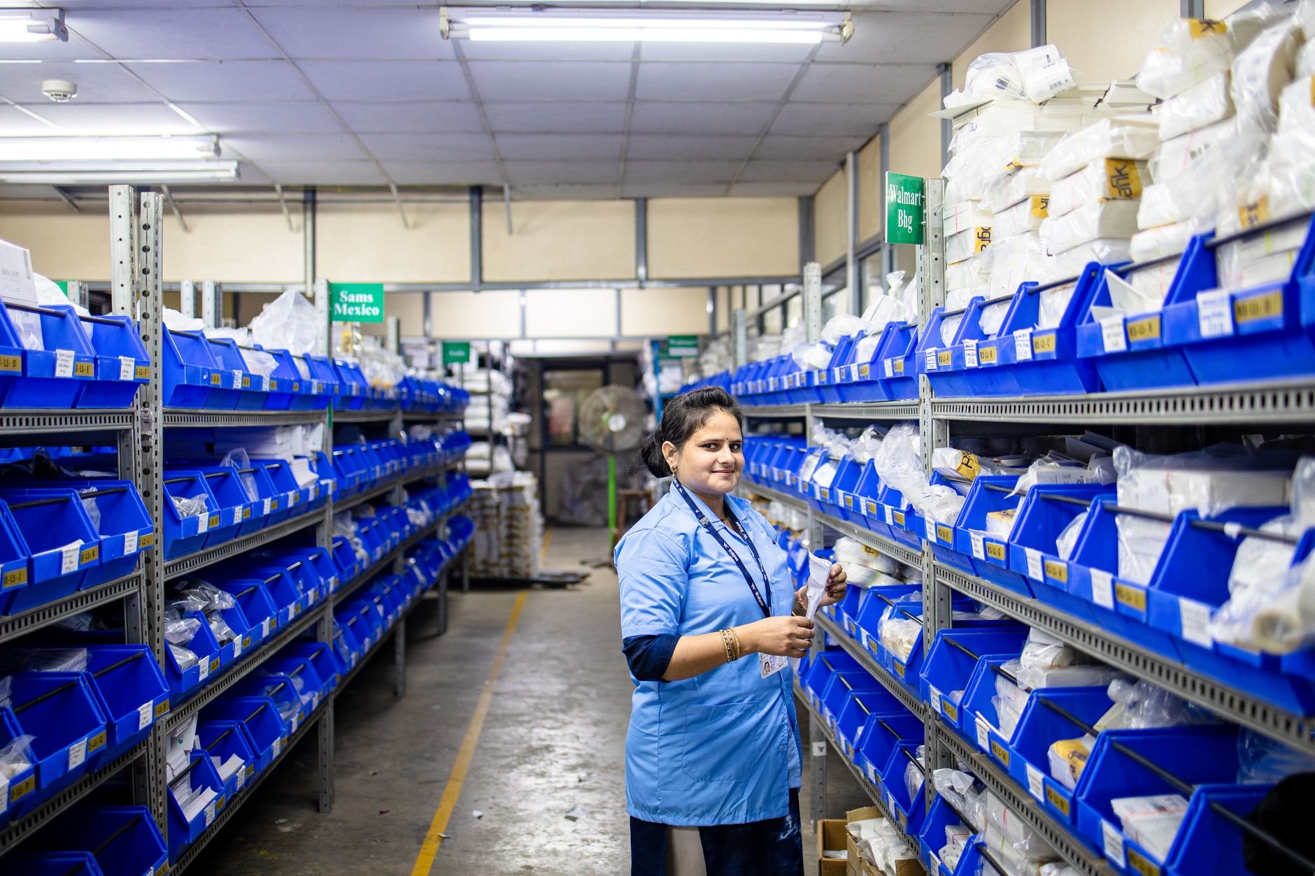 Female worker organizing inventory in a warehouse with blue bins and bright lighting.