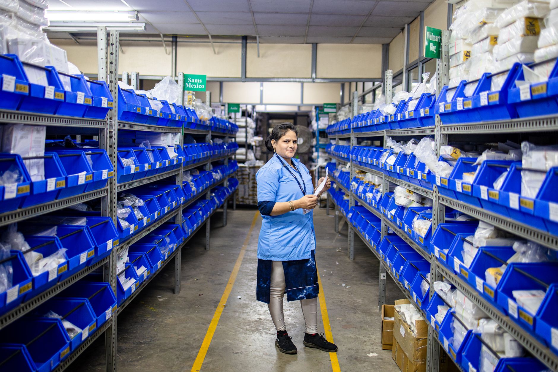 Female worker organizing inventory in a modern warehouse aisle, surrounded by blue storage bins.