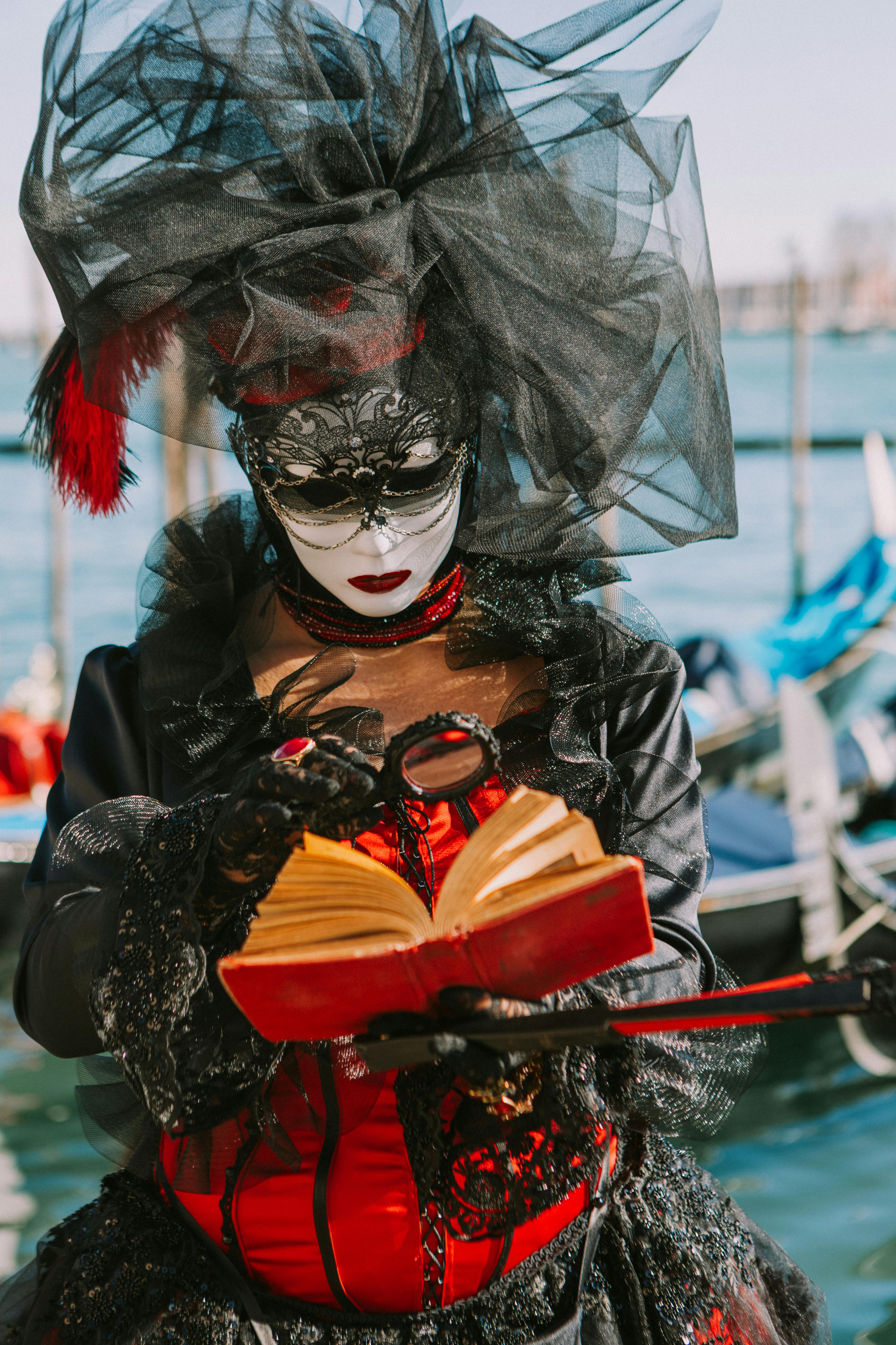 Free Masked woman in elaborate costume reads a book at Venice carnival by the water. Stock Photo