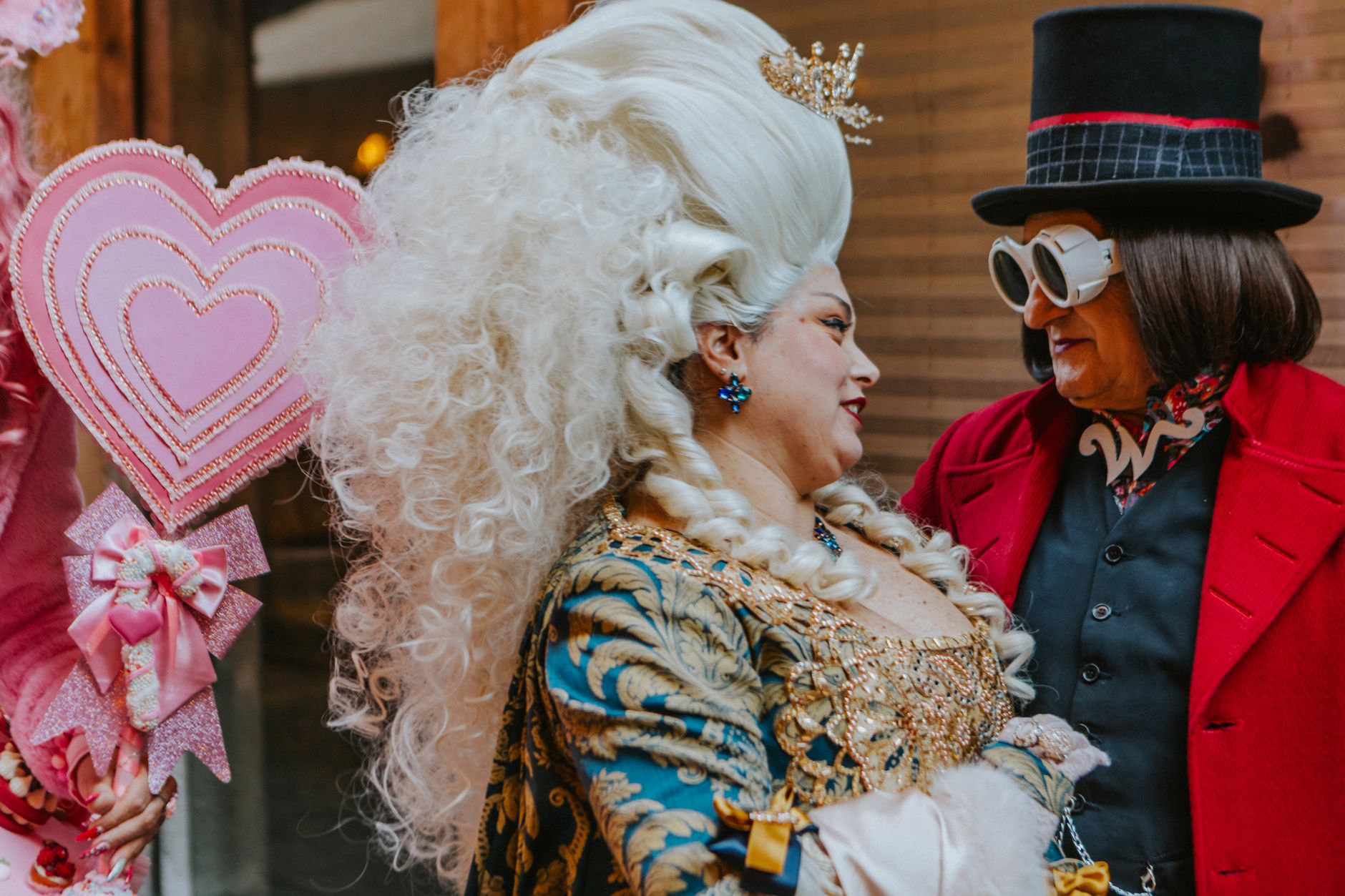 Two women in elaborate costumes at a vibrant ball, exchanging smiles with a heart-shaped decor.