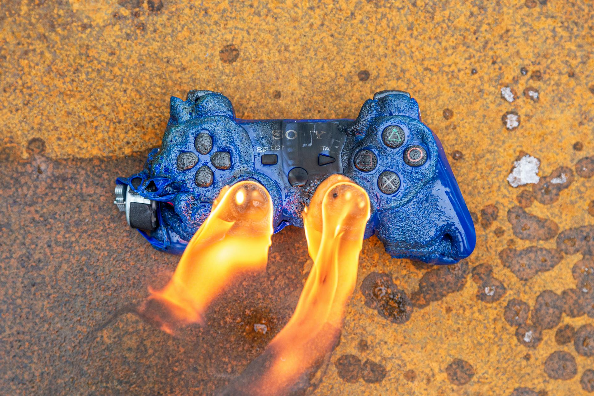 Close-up of a melting blue game controller engulfed in flames on a rusted surface.