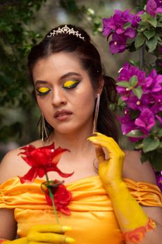 Portrait of a woman in yellow makeup and gown holding a red rose amidst vibrant flowers.