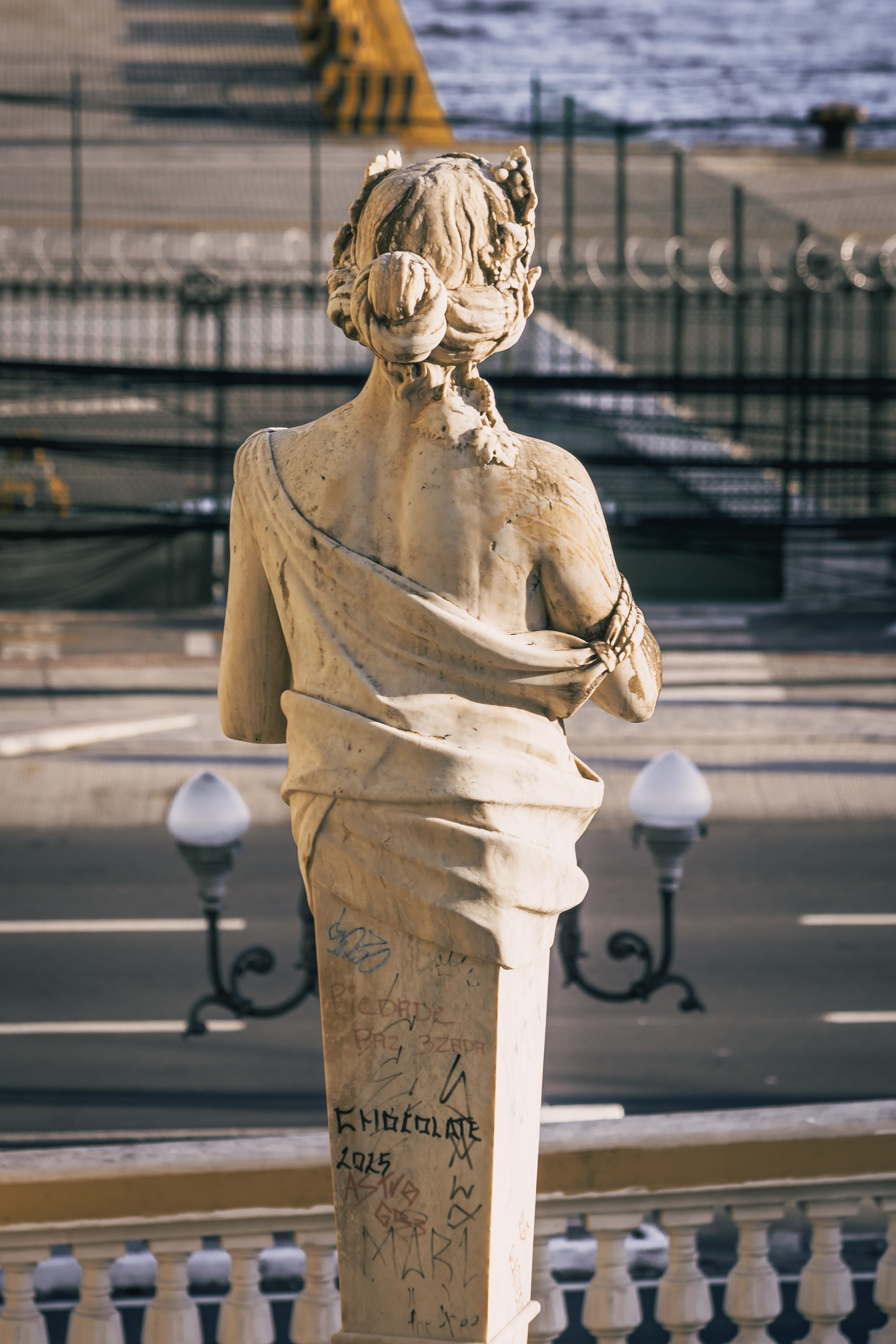 Elegant stone statue with graffiti overlooking a riverside street, embracing art within urban setting.