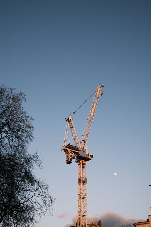 Free Tall crane and tree silhouette under a twilight sky with visible moon. Stock Photo