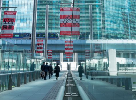 People walking on a contemporary glass bridge in an urban setting, featuring modern architecture and public transportation.