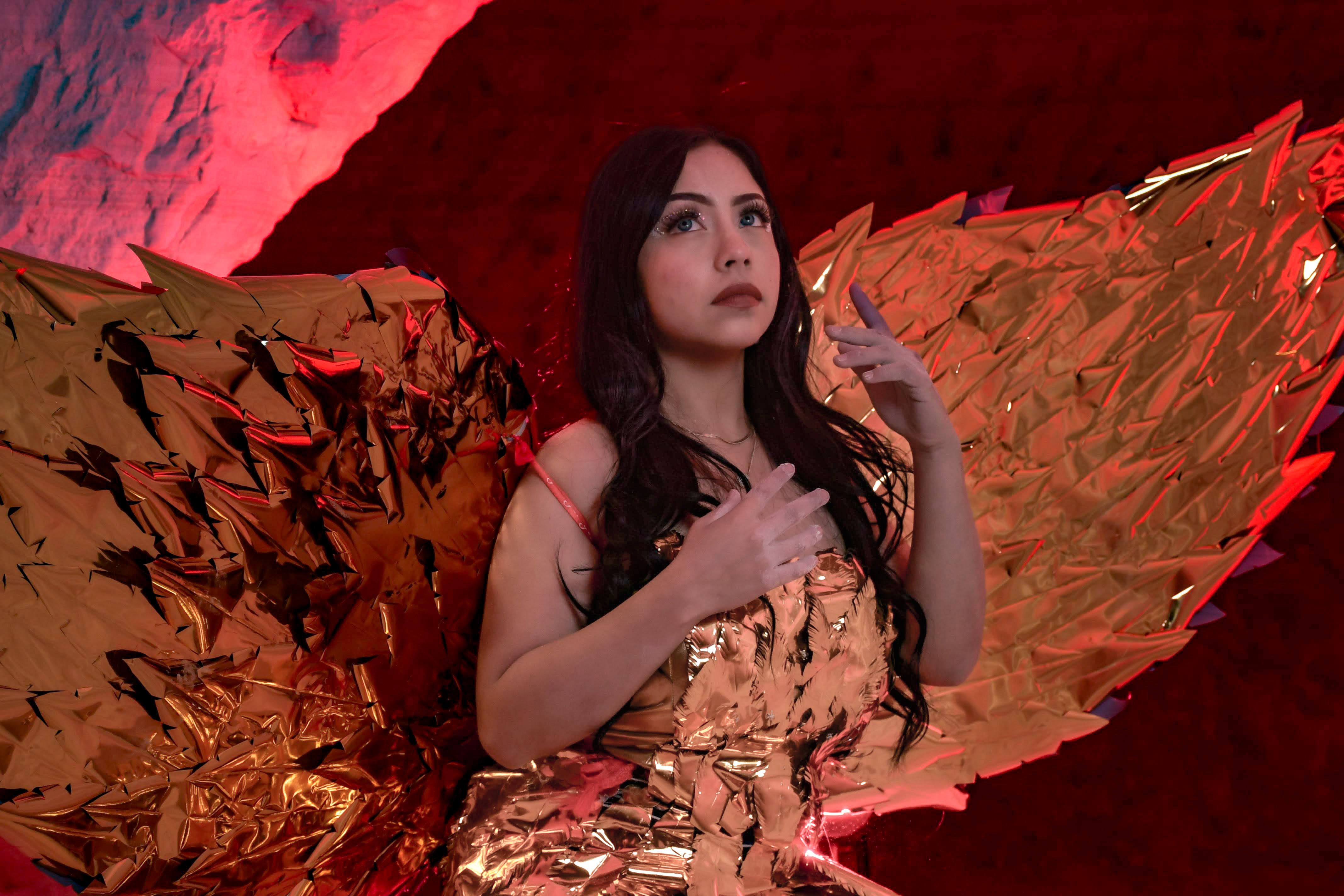 Dramatic portrait of a woman in a golden costume with wings, set against a vibrant red background.