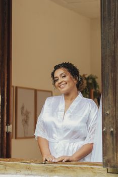 Smiling bride in a white robe leaning on a window ledge indoors.