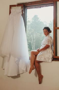 A bride in a robe sits by the window, her dress hanging beside her.