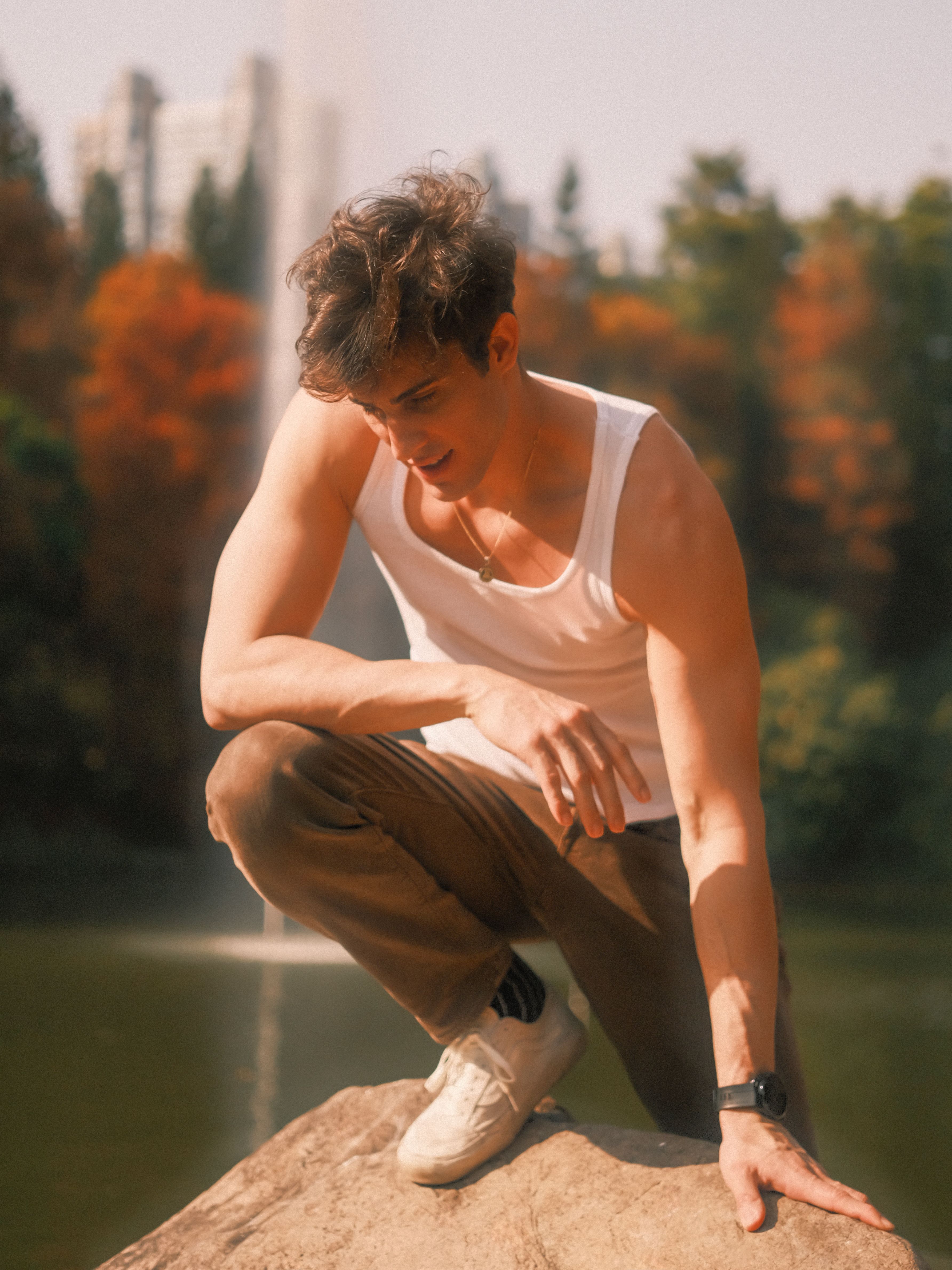 A young man in casual attire poses on a rock in an autumn park setting.