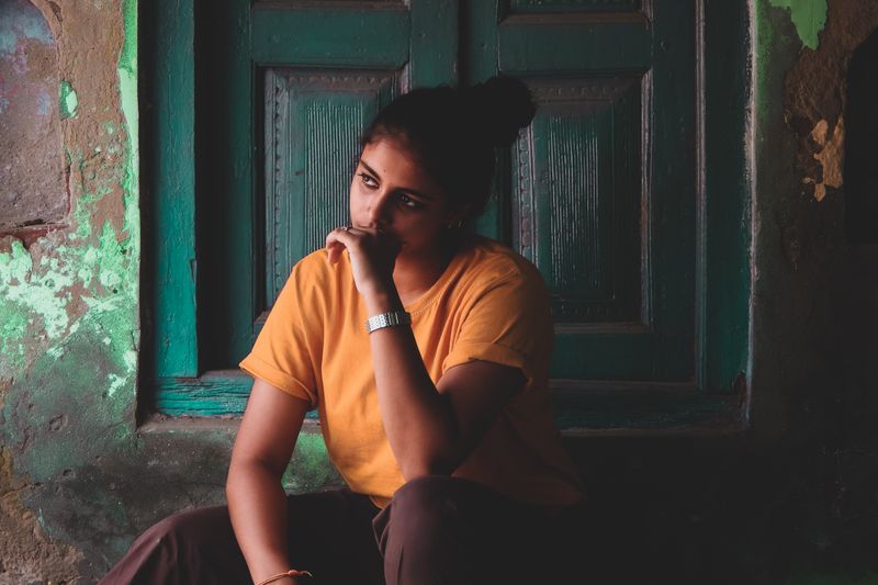 Young woman in thought, sitting by a rustic teal door, exuding a contemplative mood.