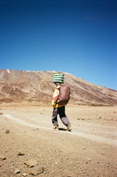 A young porter carries goods on a trek up Mount Kilimanjaro under a clear blue sky.