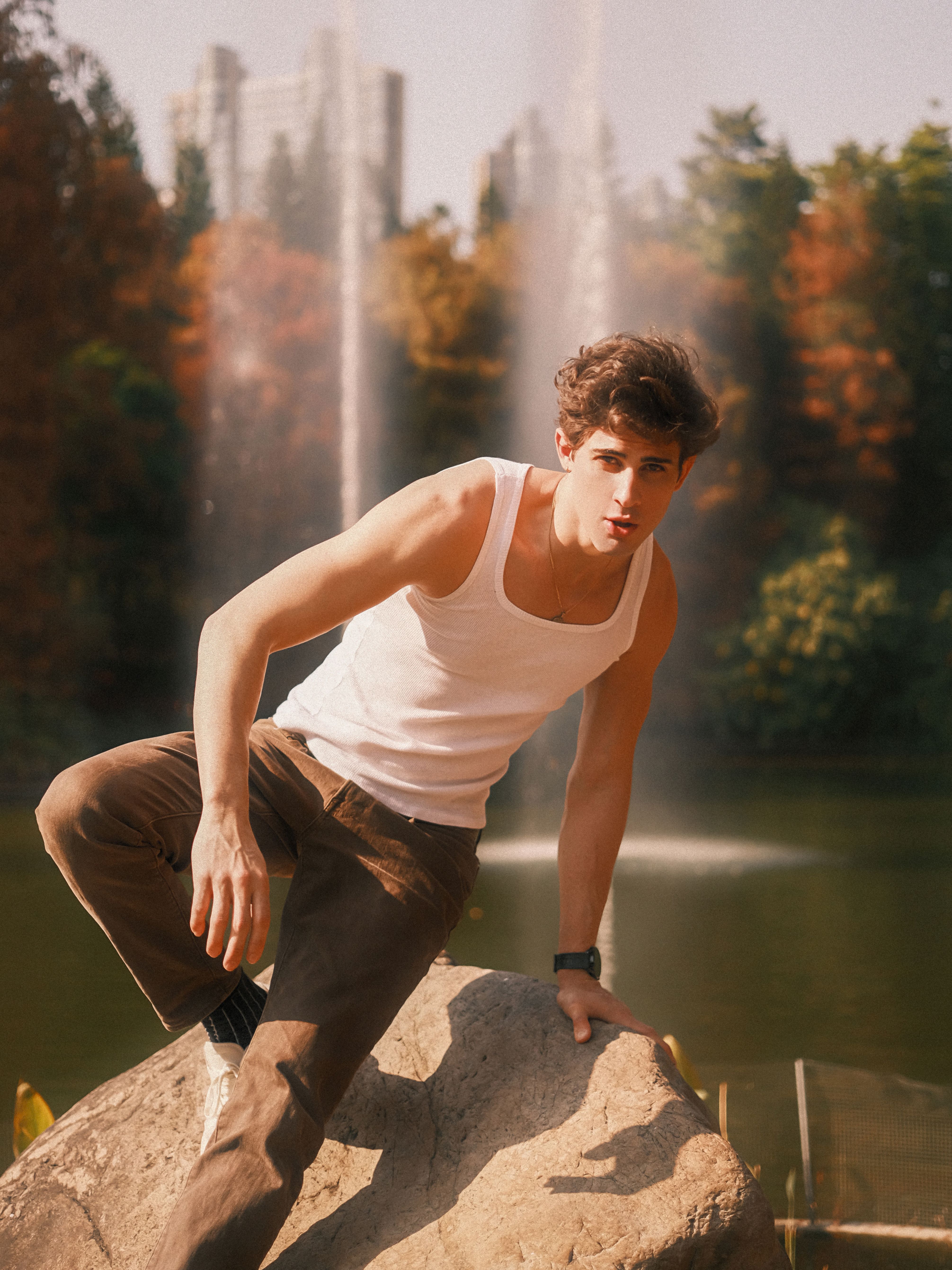 A young man in a tank top poses by a fountain in an urban park during fall.