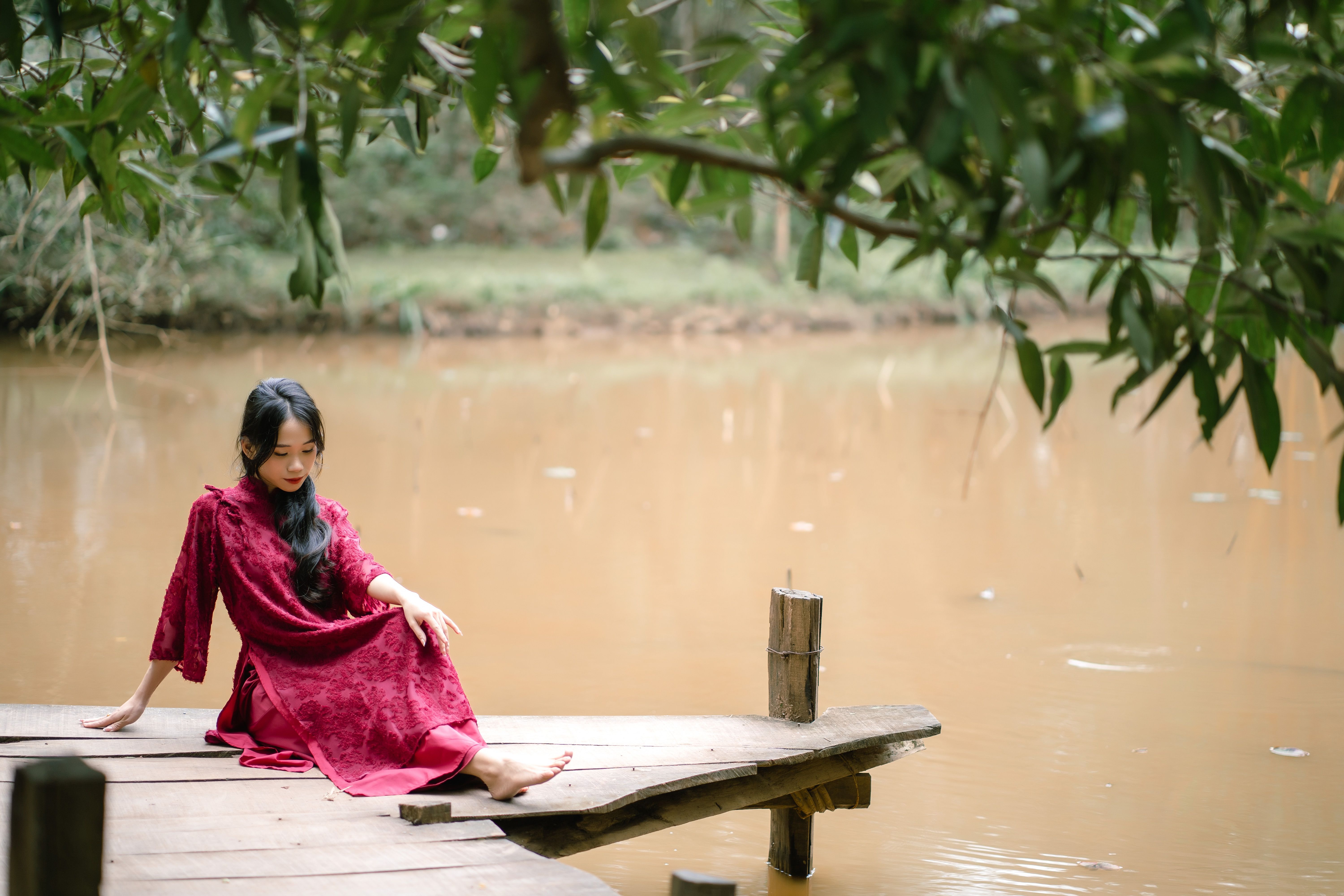 Woman in a red dress sits peacefully by a tranquil riverside setting.