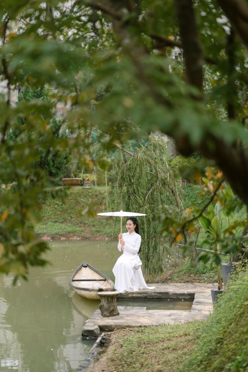 Free A serene scene of a woman in white with a parasol by a peaceful lake, surrounded by lush greenery. Stock Photo