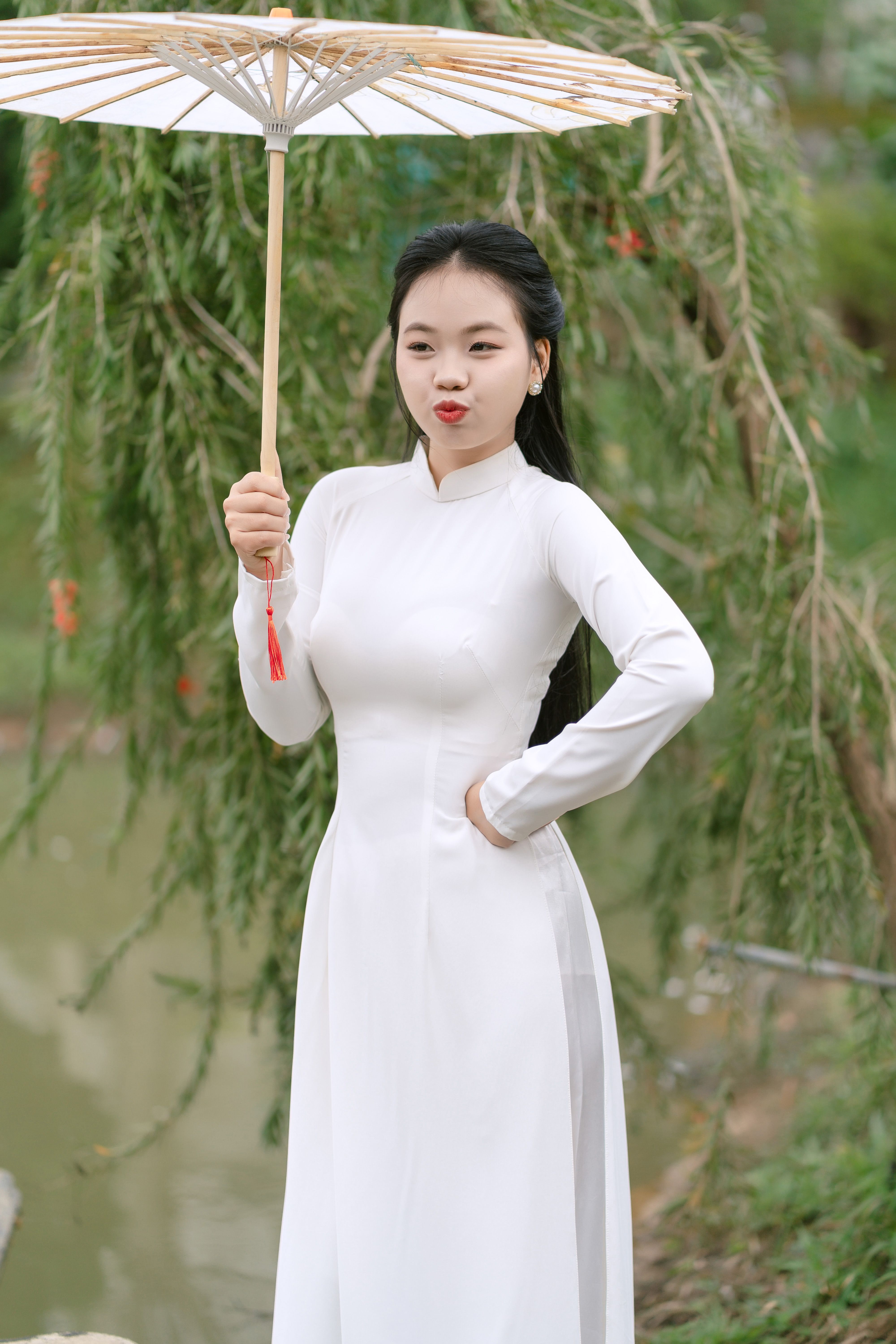 A woman in a white traditional dress holds a decorative umbrella in a serene outdoor setting.