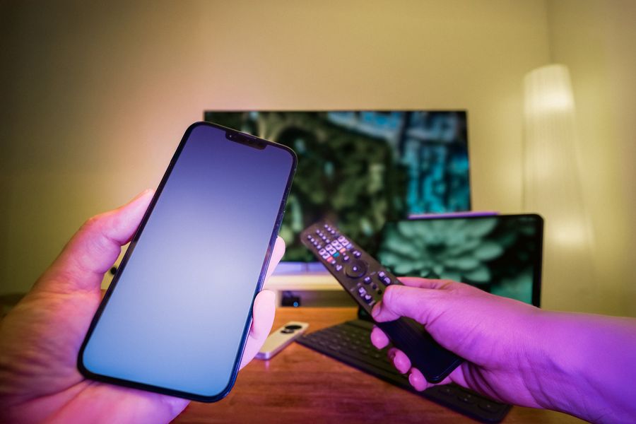 Hands holding a smartphone and a TV remote in front of a television at home