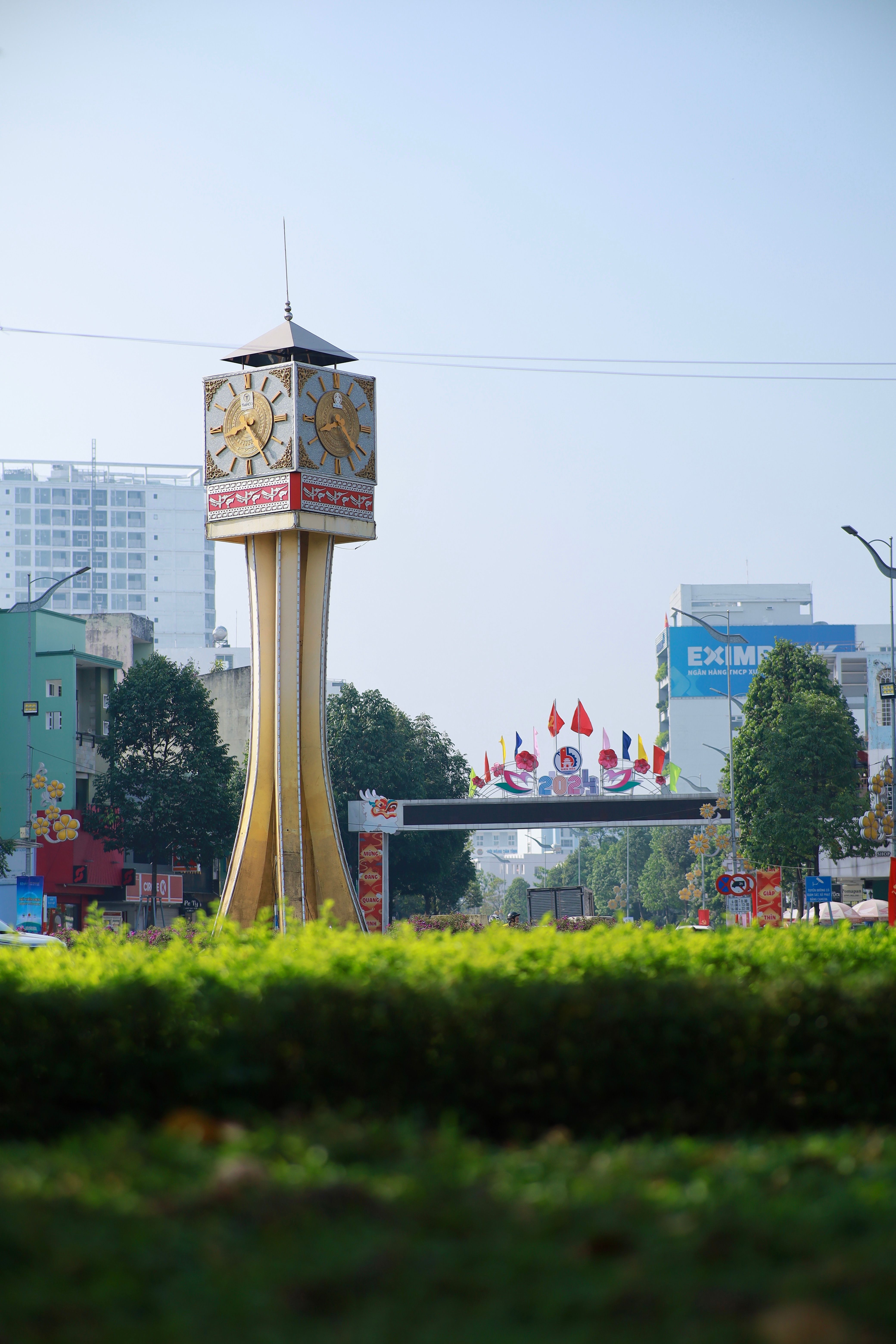A tall clock tower stands amidst city buildings and greenery on a bright day.