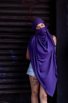 A woman wearing a purple veil stands against a dark backdrop, embodying strength and mystery in Mexico City.