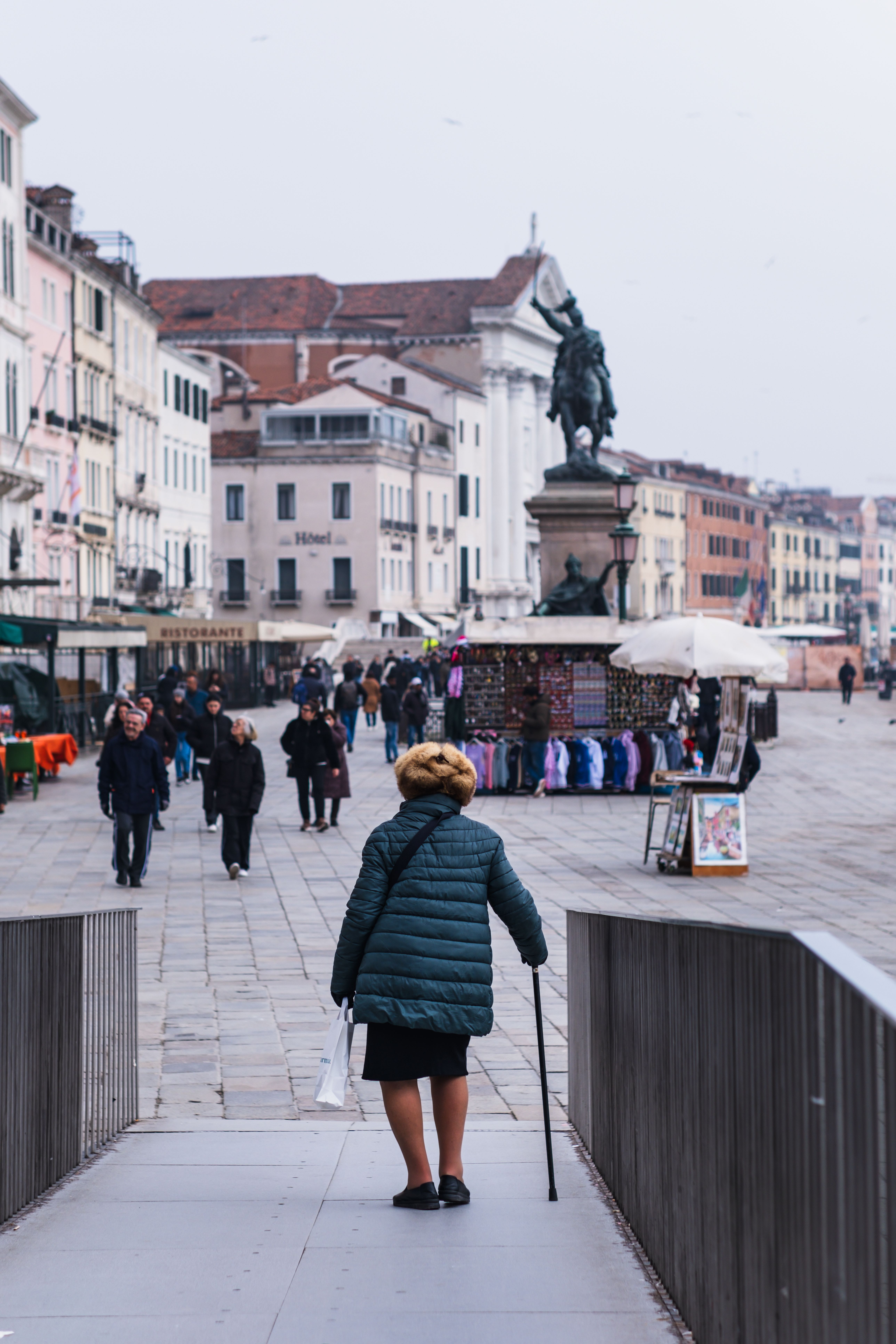 An elderly woman walks with a cane through a busy historic square in Venice, Italy.